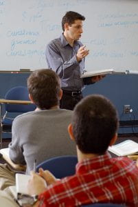 A male professor is teaching in a classroom in front of a whiteboard covered in writing. Two male students sitting in desks have their backs turned to the camera and are watching the professor.