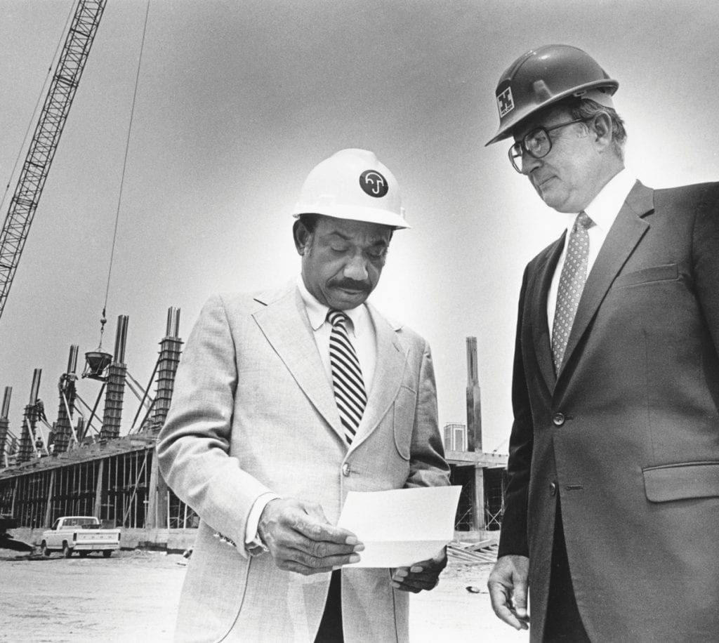 Herman Russell in suit and hard hat looking at a document along with developer Robert Holder, also in suit and hard hat. A building is under construction behind them, with pillars of concrete and rebar extending skyward and a crane to the side. (Photo Courtesy the Atlanta Journal-Constitution via the New Georgia Encyclopedia)