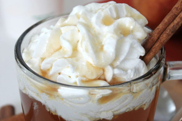 A glass mug of caramel apple cider with whipped cream on top and a cinnamon stick garnish