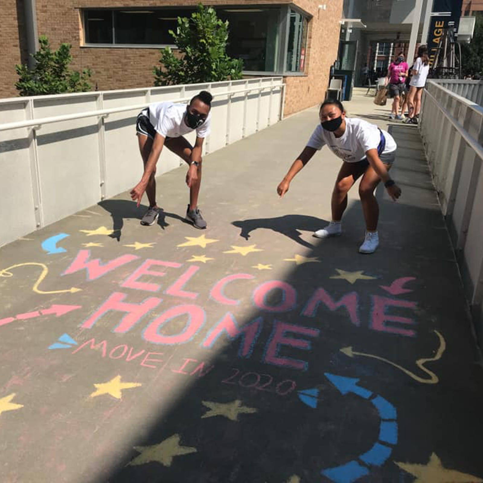 Two students pointing to 'Welcome Home' chalked on the ground.