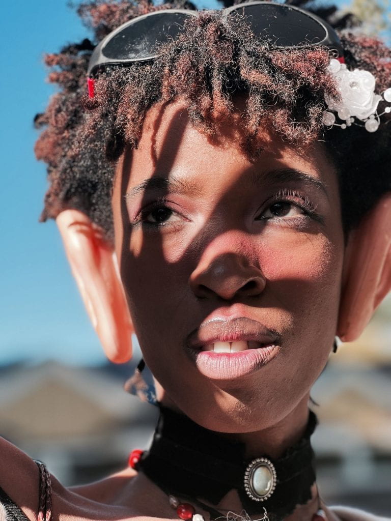 Adia is outdoors with a blurred background, wearing extra large ears and a decorative white flower in hair.