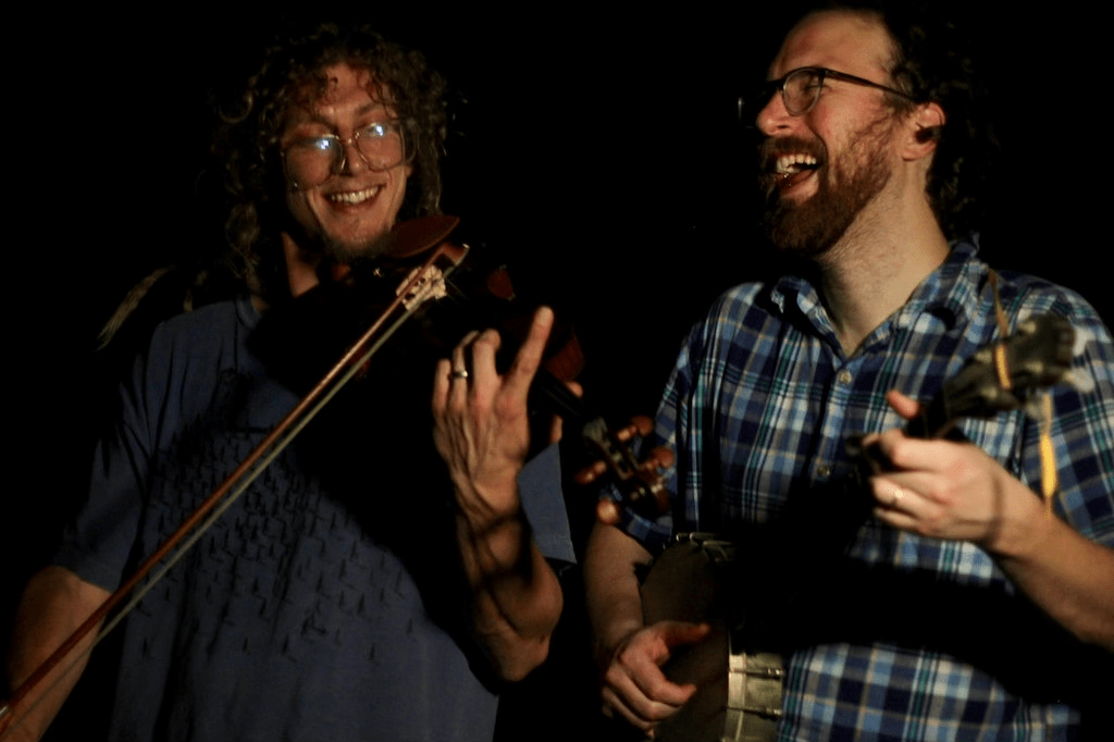 Majid (left) plays violine and Ben (right) plays banjo. The background is completely dark. They are smiling or laughing.